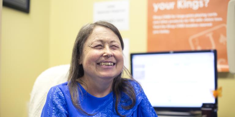 Woman sitting in a chair, smiling. Behind her is a computer screen and an orange poster on a yellow wall.