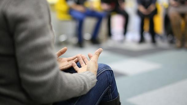 A person sits in a group circle. Their hands are clasped in their lap.
