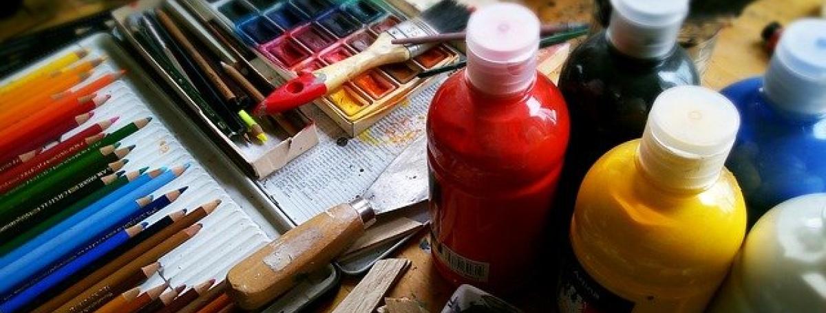 A tabletop covered in art supplies; coloured pencils, paintbrushes and jugs of paint.