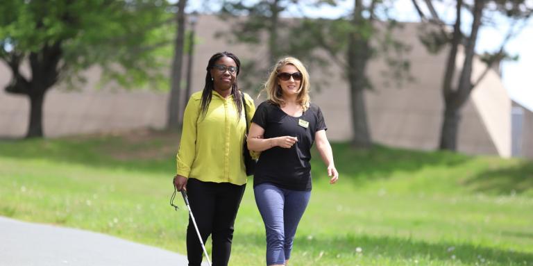 A woman provides sighted guide to another woman holding a white cane