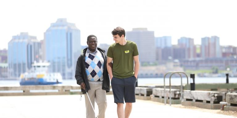 A man with a white cane walks on the Dartmouth waterfront guided by a younger man