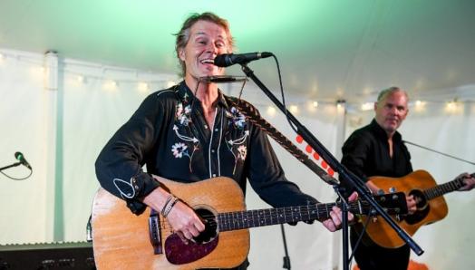 Jim Cuddy se produit en direct au centre Lake Joe d'INCA. Il est sur scène, joue de la guitare et chante au micro. Un autre membre de son groupe joue de la guitare en arrière-plan.