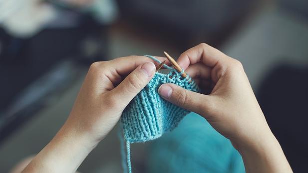 Two hands hold knitting needles and knit a blue fabric.