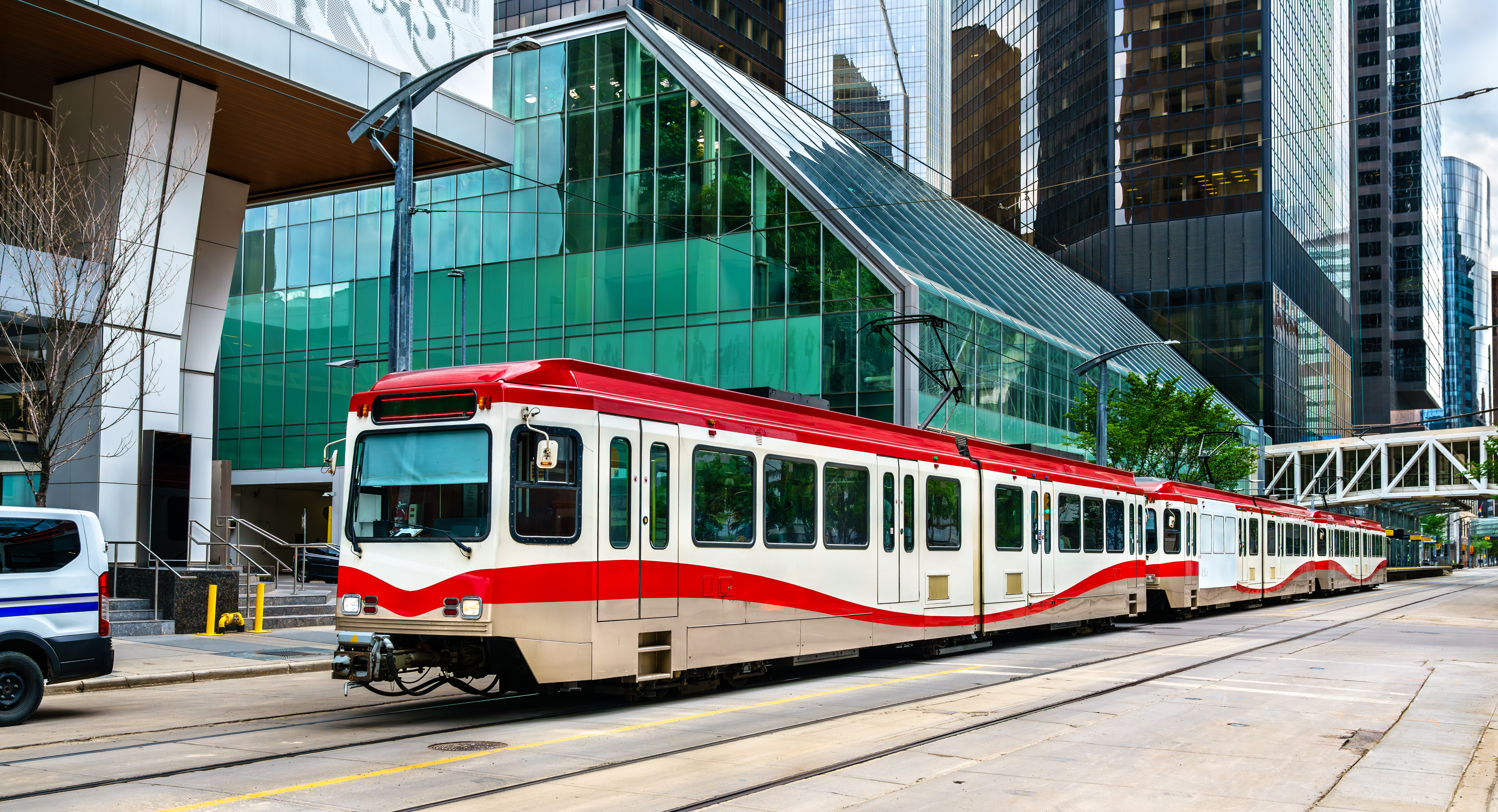 A red and white CTrain car glides along an outdoor light rail track in downtown Calgary. In the centre of the image is a brushstroke overlay with the text: Get on Board!