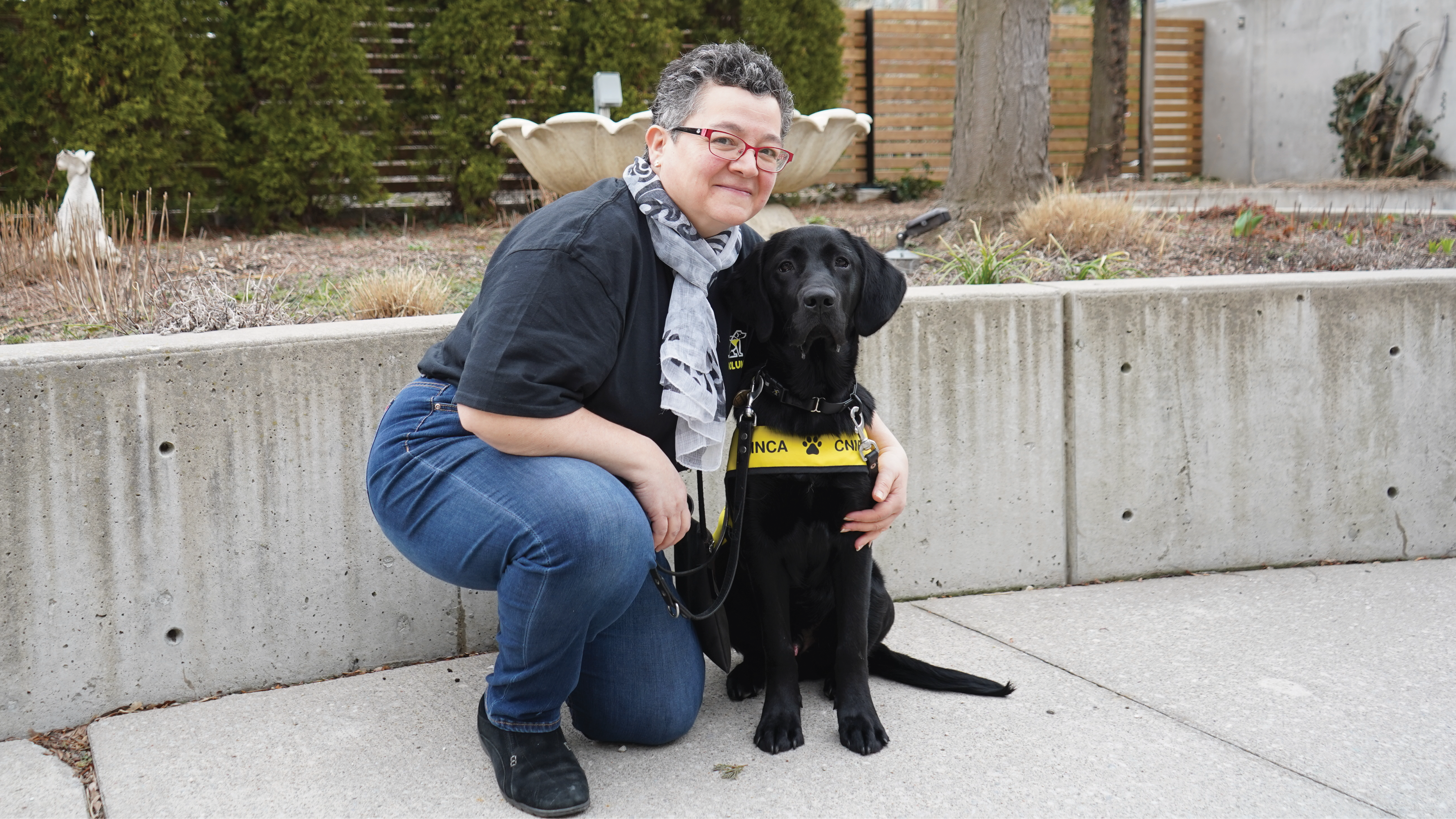 Outdoors, a puppy raising volunteer crouches down on the ground next to a black puppy in training. The puppy raiser has her arm around the pup and the pup is wearing a yellow "future guide dog in training" vest. 