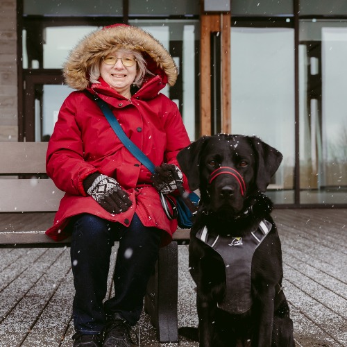 Cheri smiles and sits on a bench, and her guide dog, Sassy, sits on the pavement to her right. Sassy is a black dog in harness. It’s lightly snowing, and Cheri is wearing a red winter jacket and gloves. 