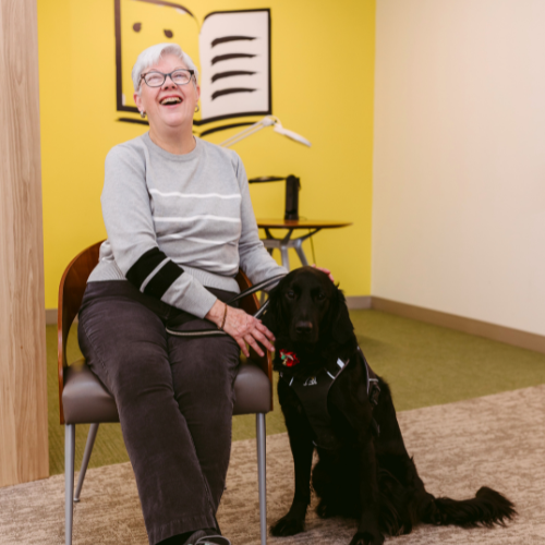 At a CNIB office, Penny sits in a chair. Her guide dog, Honour, a black Labrador retriever-golden retriever cross, sits at her feet. Penny pets Honours head and smiles with joy.