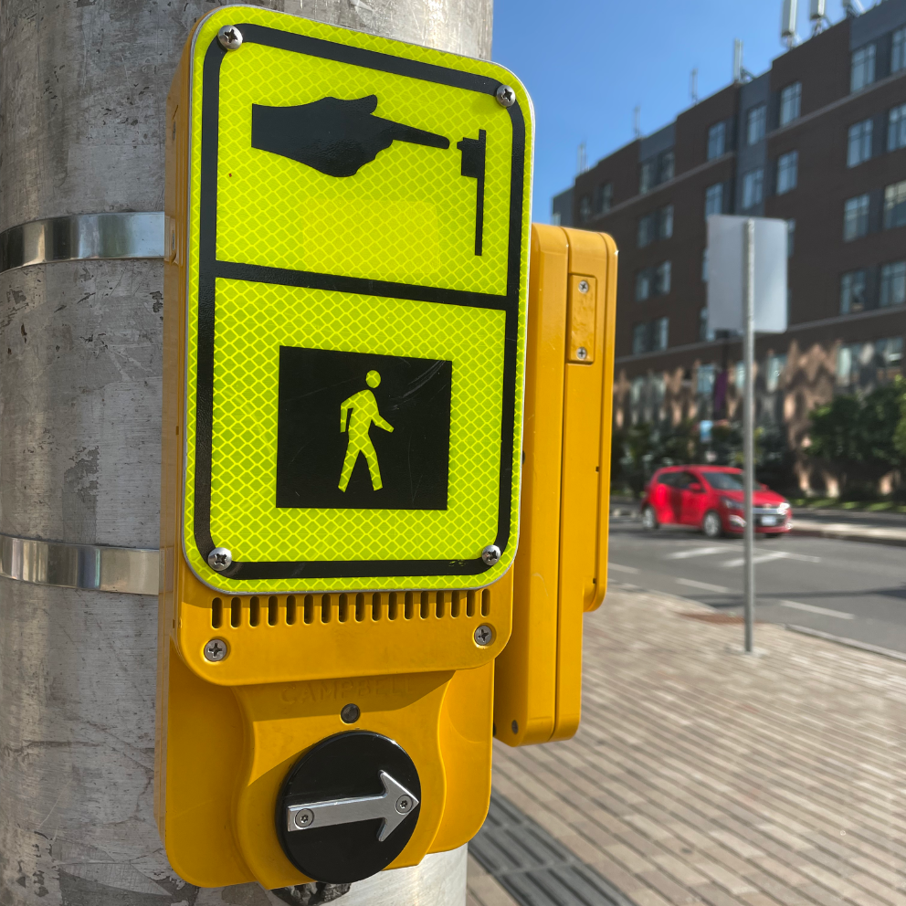 An Accessible Pedestrian Signal (APS) mounted on a metallic pole at a crosswalk in Ottawa. The surrounding area shows an urban environment with a brick-paved sidewalk, a red car in the background, and modern buildings under a clear blue sky.