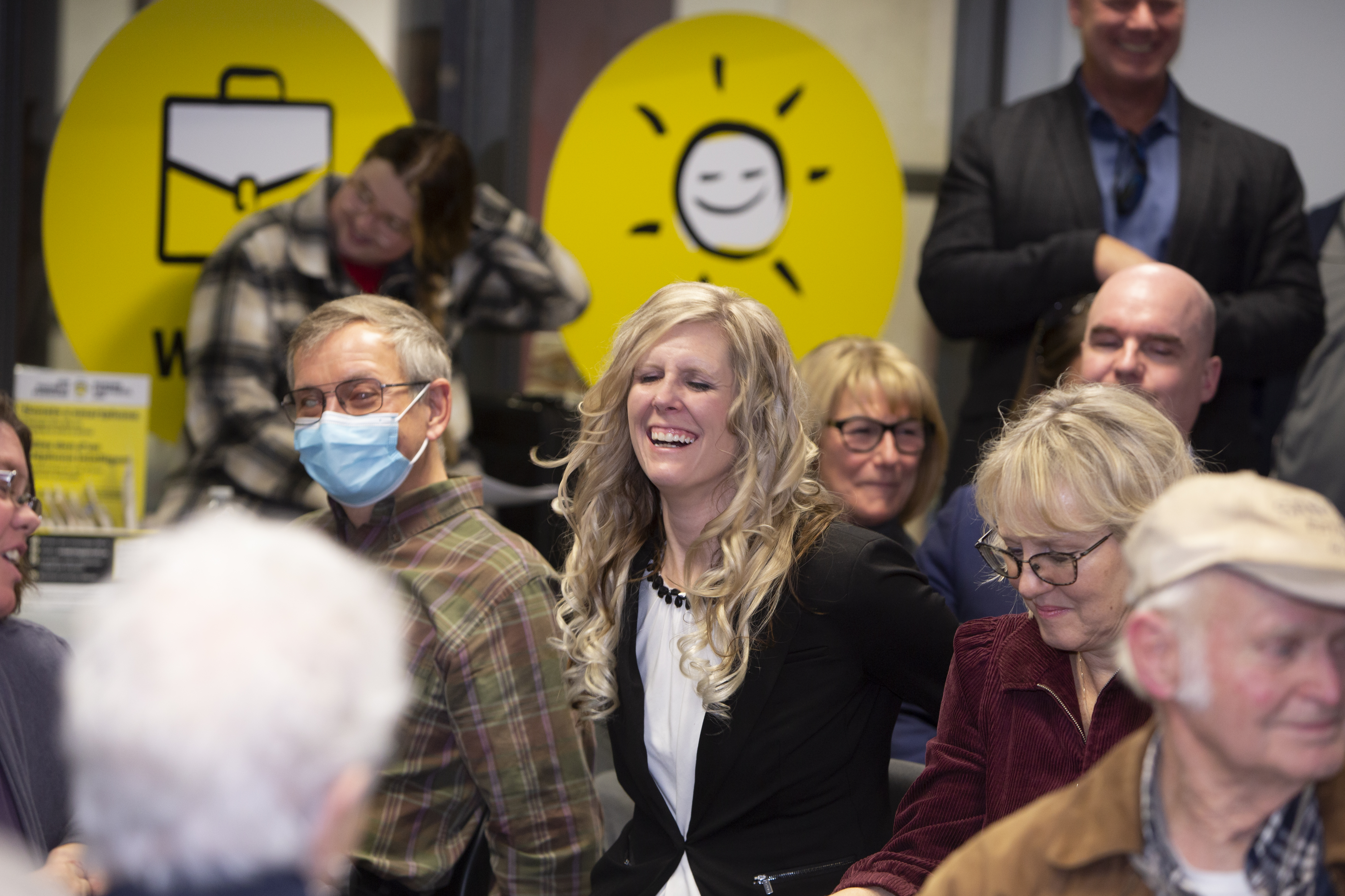 a group of people smile and listen as a crowd. In the centre, a woman with blond hair has a huge smile on her face.