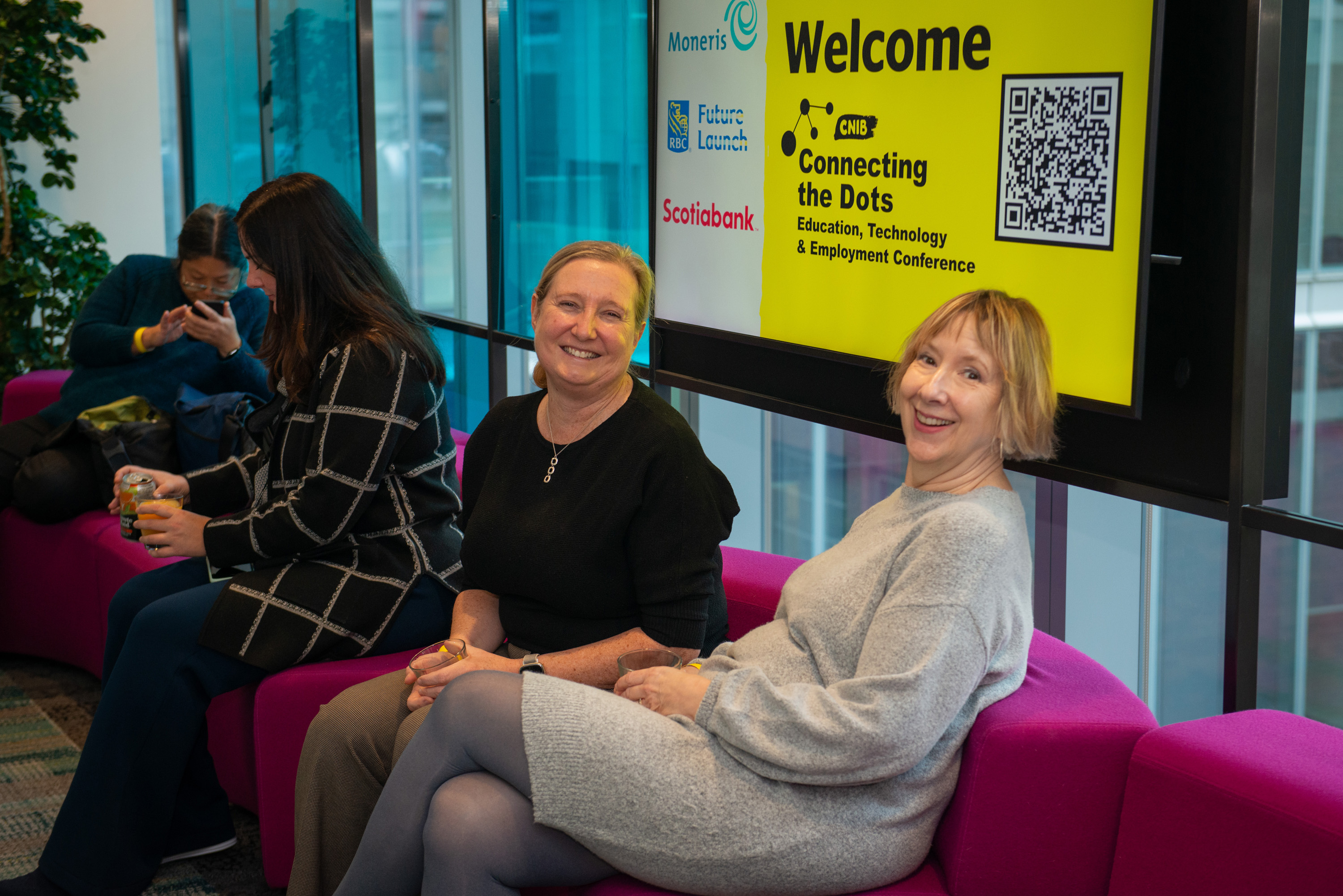 Two women sit on a couch with a Connecting the Dots sign behind them. 