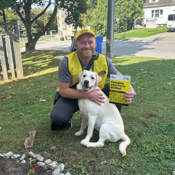 A canvasser working on behalf of CNIB kneels beside a dog in a grassy front yard. The canvasser wears a yellow CNIB hat and vest and holds up a large card featuring facts about blindness.