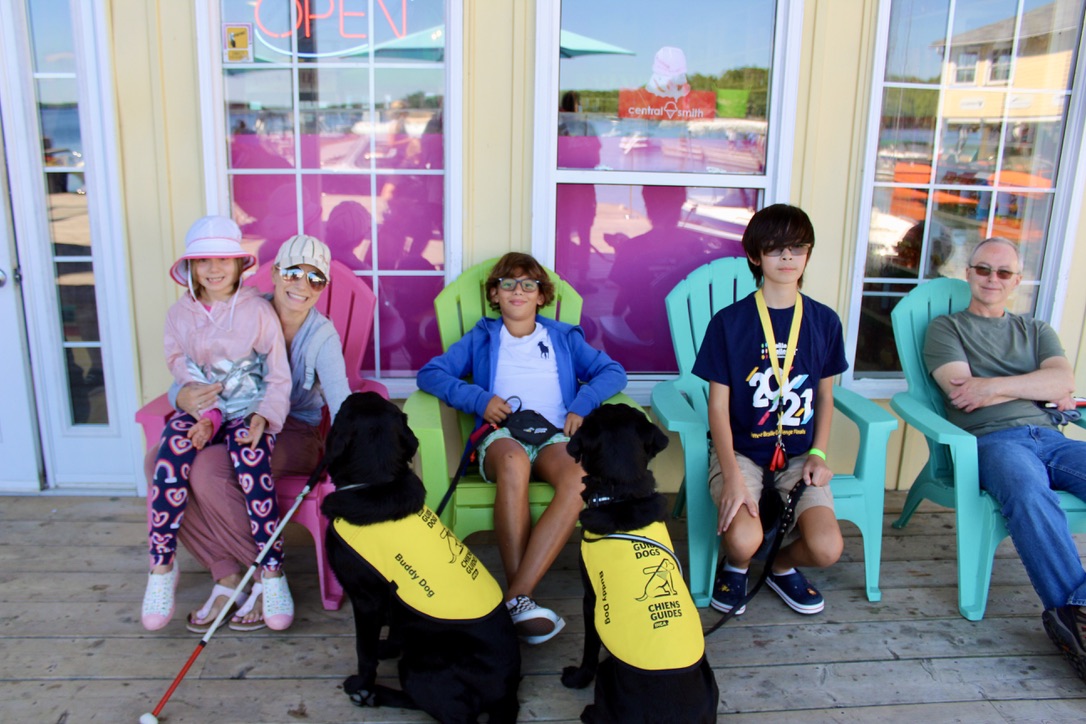 Gabriel, 12 ans, est assis dans une chaise Muskoka colorée, détendu et souriant, entouré d’autres familles et campeurs heureux du Centre Lake Joe d’INCA et de deux chiens compagnons d’INCA portant des gilets jaunes.