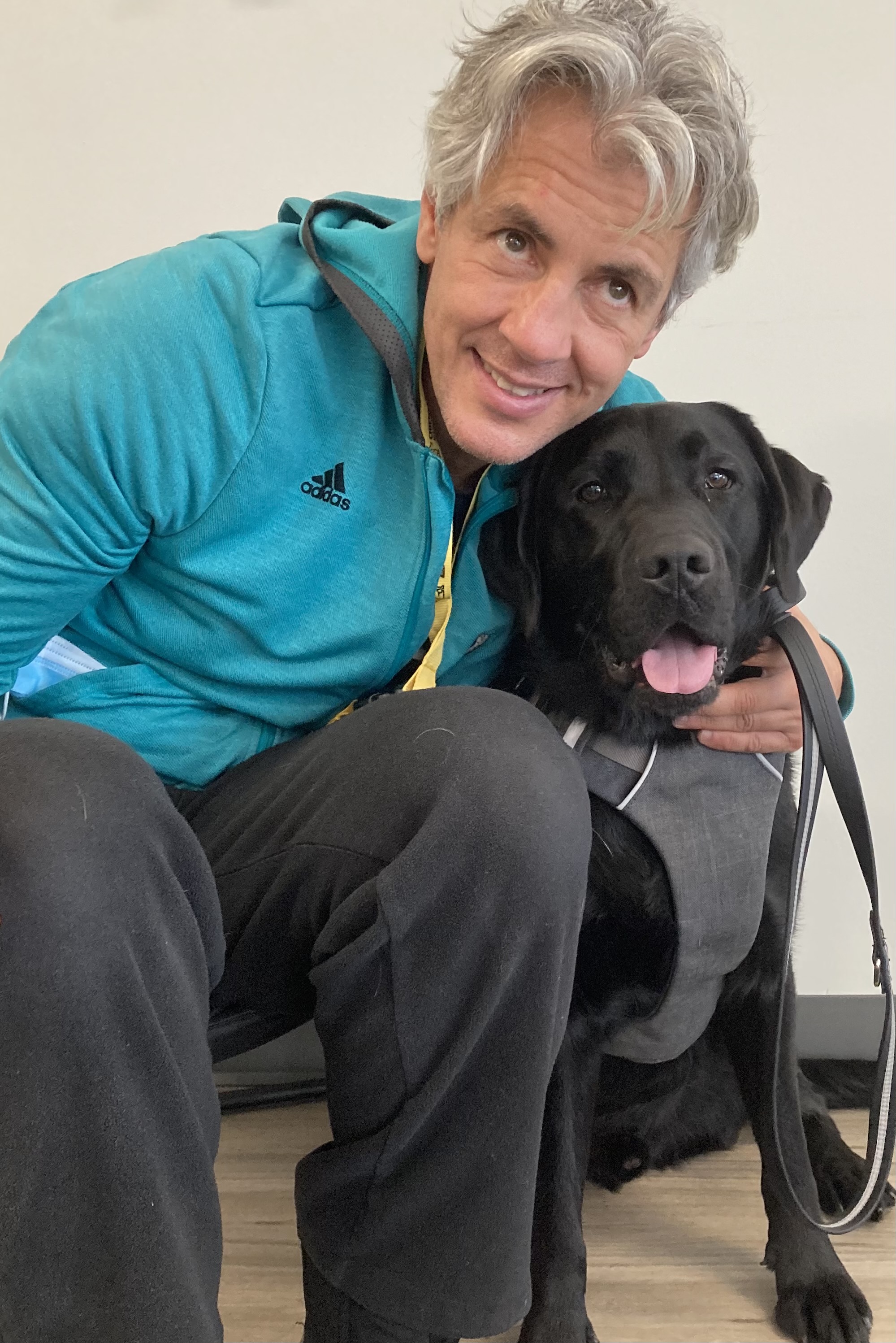 A picture of Ary wearing a turquoise Adidas jacket and black pants, sitting on the floor giving Don, a CNIB Guide Dog, a belly rub while smiling for the camera. To the right of the photo in white space, text reads: Humans with CNIB Guide Dogs – Ary & Don