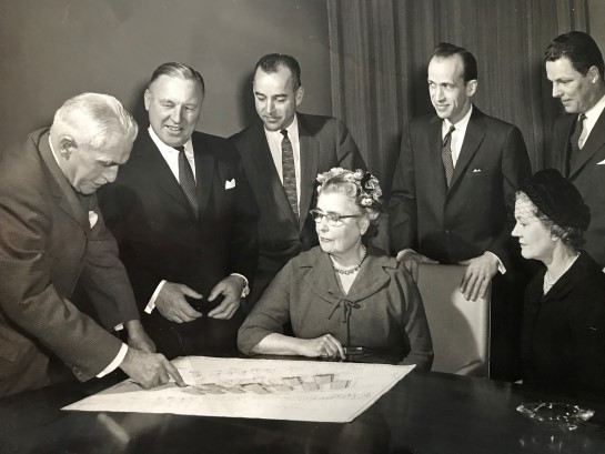 An old black & white photograph of the Lake Joseph Building-Planning Committee studying pans for the Adjustment-Training and Holiday Centre (Seated: Elsinore Burns and Margot Brechenridge. Standing left to right: Wilfred James (Chairman), Norman Long, J.A Peguegnat, J. Mossop, J.A. Clark).