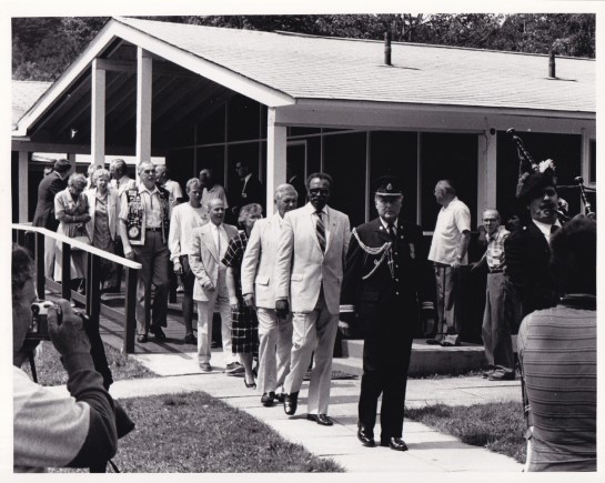 A black & white photograph of Lincoln Alexander, Lieutenant Governor of Ontario, following bagpiper and man in uniform at CNIB Lake Joe, July 20, 1986.