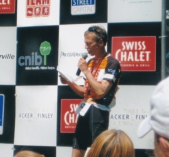 A photograph of Joe Finley at CNIB Lake Joe. He is holding a microphone and standing in front of a branded step and repeat. 2008