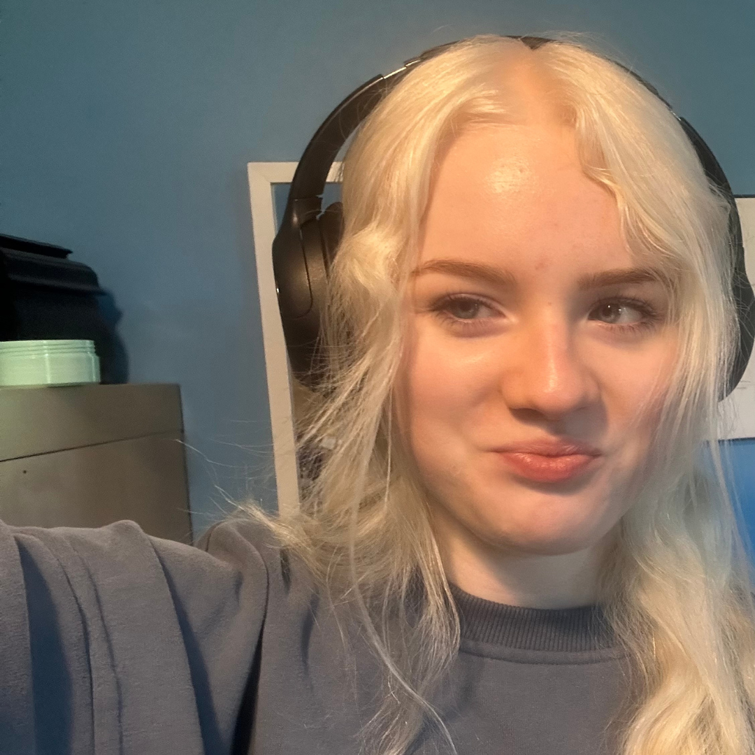 A selfie-style photograph of Acacia. Acacia has long platinum white hair and is wearing headphones over he ears and a long grey sweatshirt.