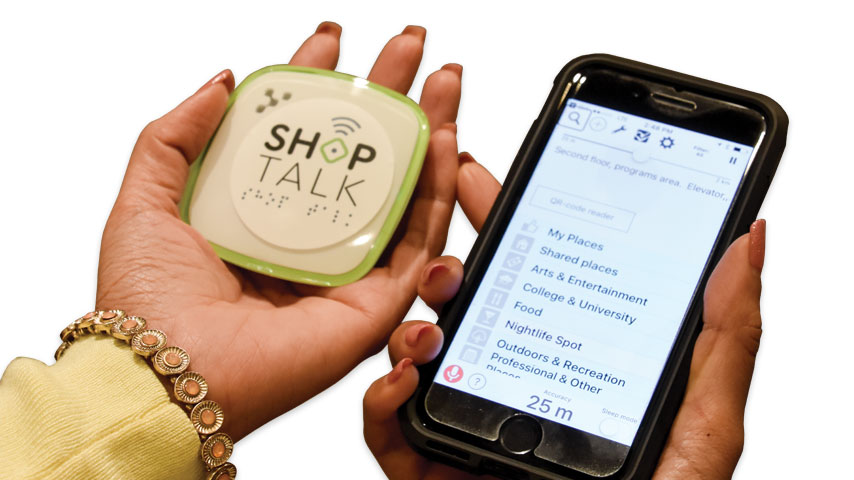 Photo d'une main de femme tenant un petit appareil de connexion à  BlindSquare zur lequel est marqué Shop talk et un téléphone cellulaire dans l'autre.