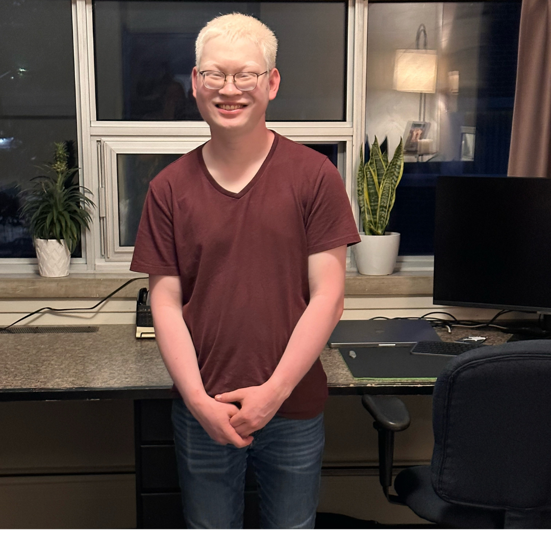 Adian stands in-front of a home office/computer workstation. He is smiling and standing with his hands clasped together. Aidan has platinum white hair, wears glasses and a brown t-shirt with jeans.