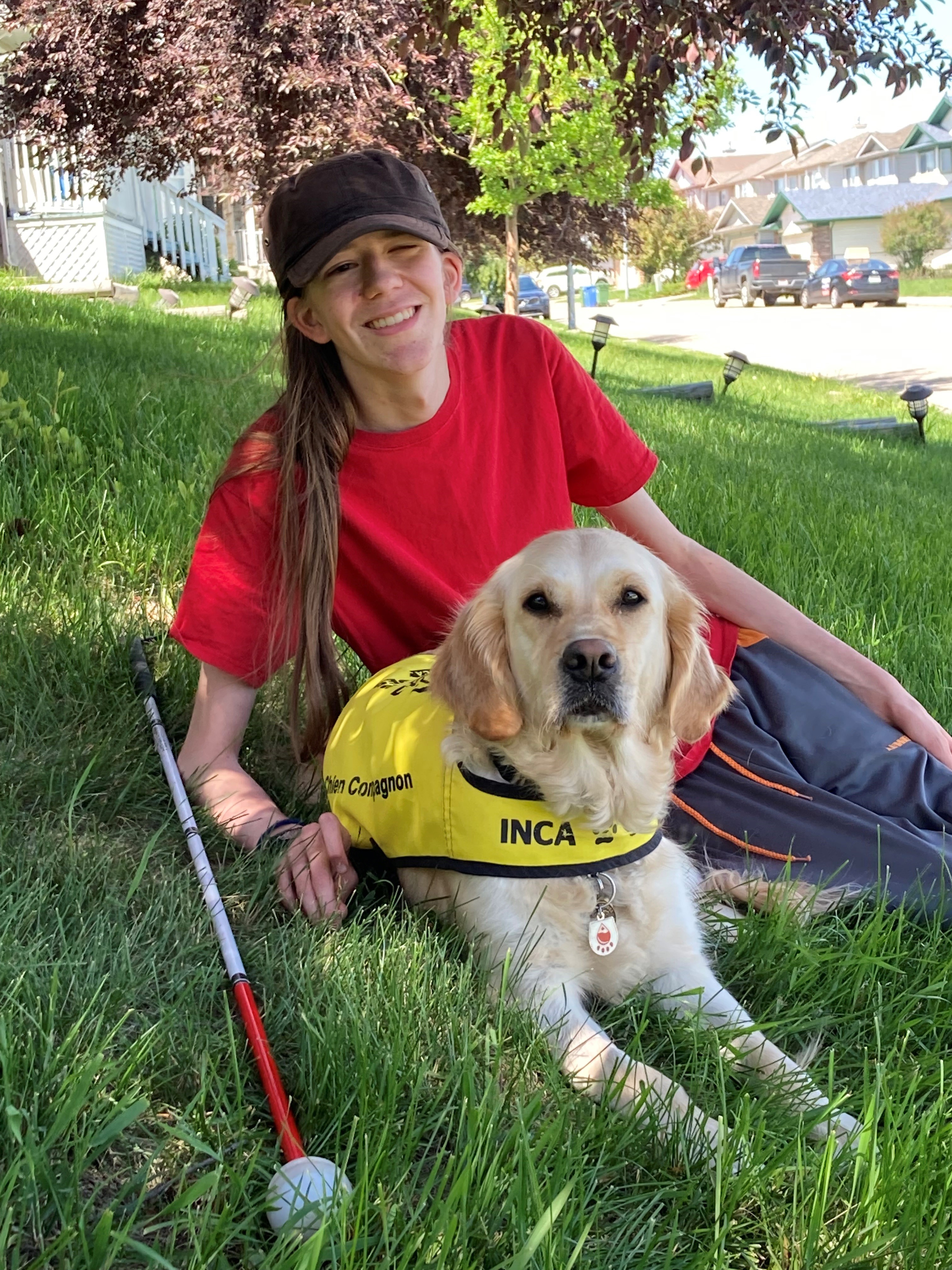 Zach reclining on the grass with his buddy dog Elsie, a golden retriever who is wearing a yellow CNIB buddy dog vest. Zach’s white cane sits on the grass next to him.
