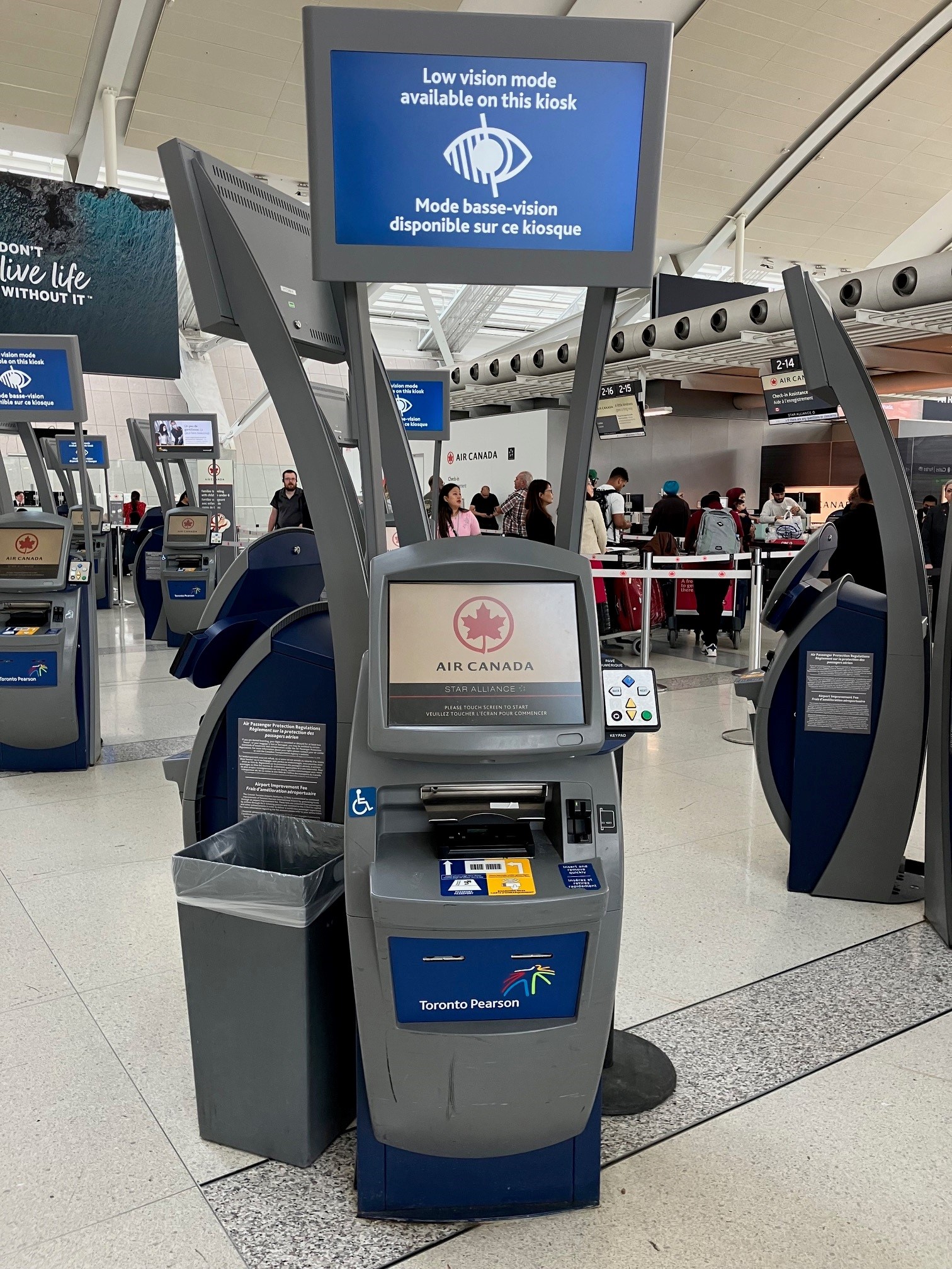 A self-service check-in kiosk at Pearson Airport. The kiosk shows an Air Canada logo on the screen with a sign mounted above that reads, "Low vision mode available on this kiosk." A panel with accessible navigation buttons is located on the right side of the kiosk, next to the touchscreen.