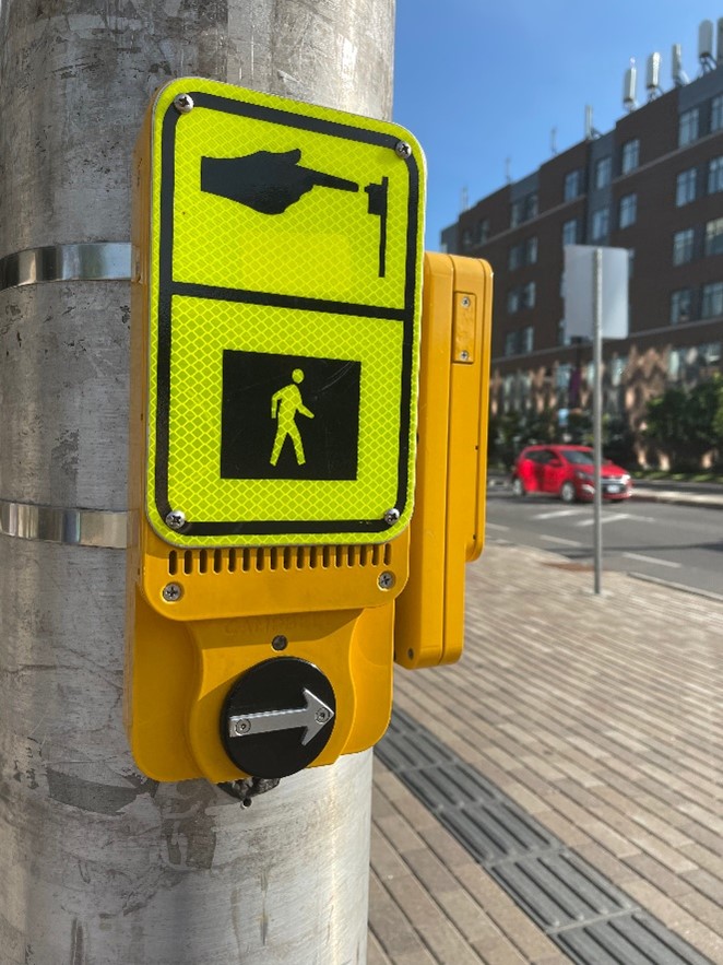 An Accessible Pedestrian Signal (APS) mounted on a metallic pole at a crosswalk in Ottawa. The surrounding area shows an urban environment with a brick-paved sidewalk, a red car in the background, and modern buildings under a clear blue sky.