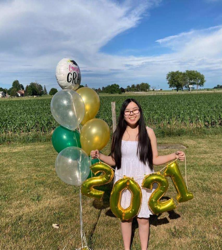 Stormy stands in front of a rural corn field on a sunny summer day. She wears a sundress and is holding a bouquet of graduation-themed balloons