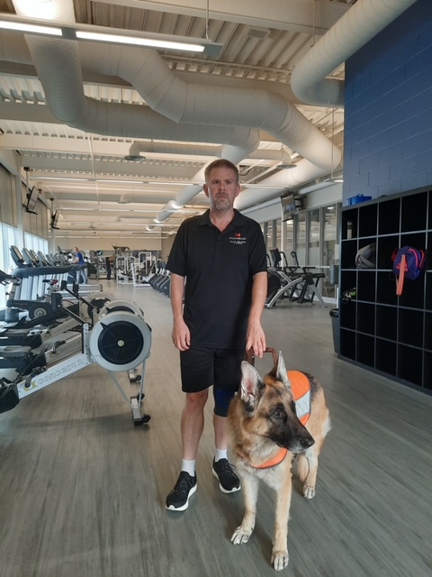 Bob et son chien-guide Berger allemand à l’intérieur d’un gymnase. La salle de gym est remplie d’équipement d’entraînement.