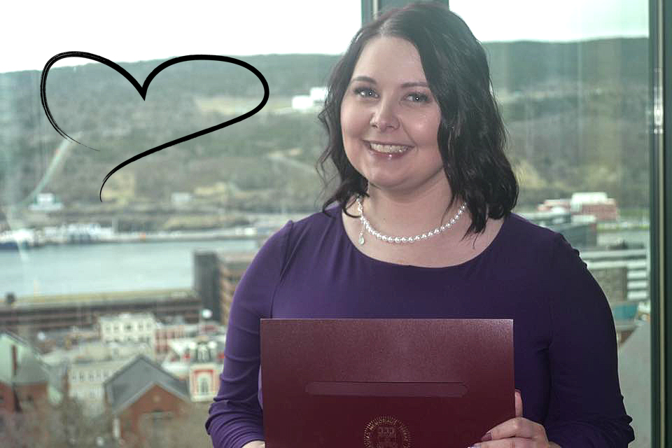 Brittany Farrell, smiling for the camera, standing in front of a window that overlooks the city of St. John’s, Newfoundland. A heart graphic can be seen against the window.