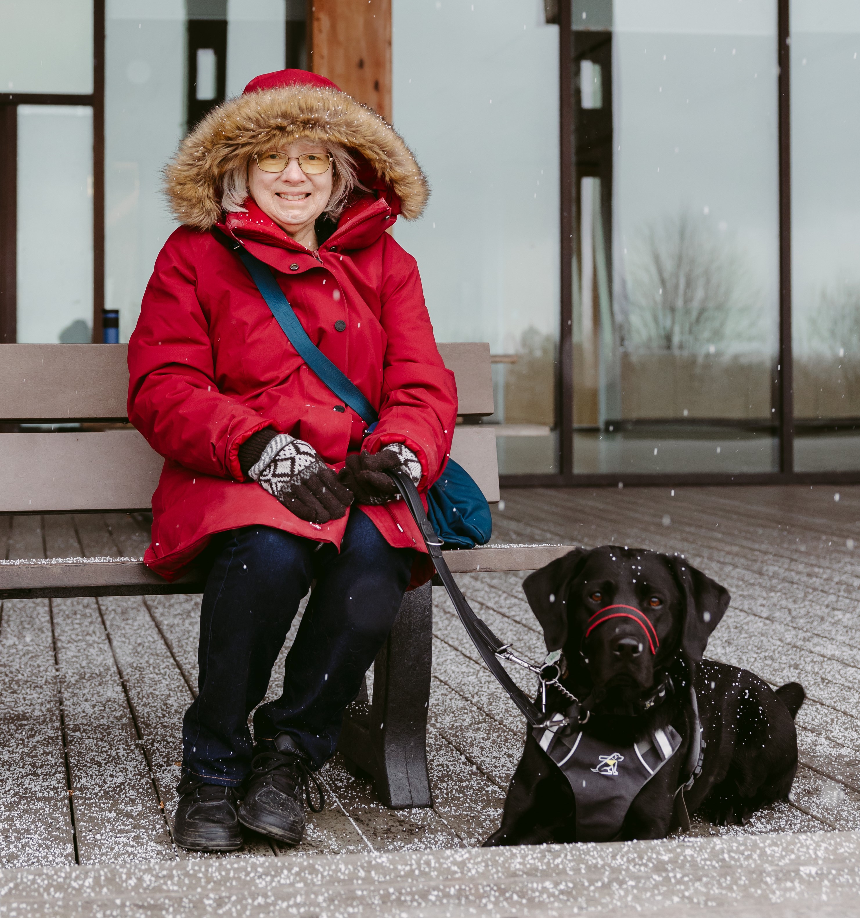 Cheri, who wears a hooded red parka, sits on a wooden bench with Sassy, her CNIB black Labrador guide dog, by her side on a snowy day.