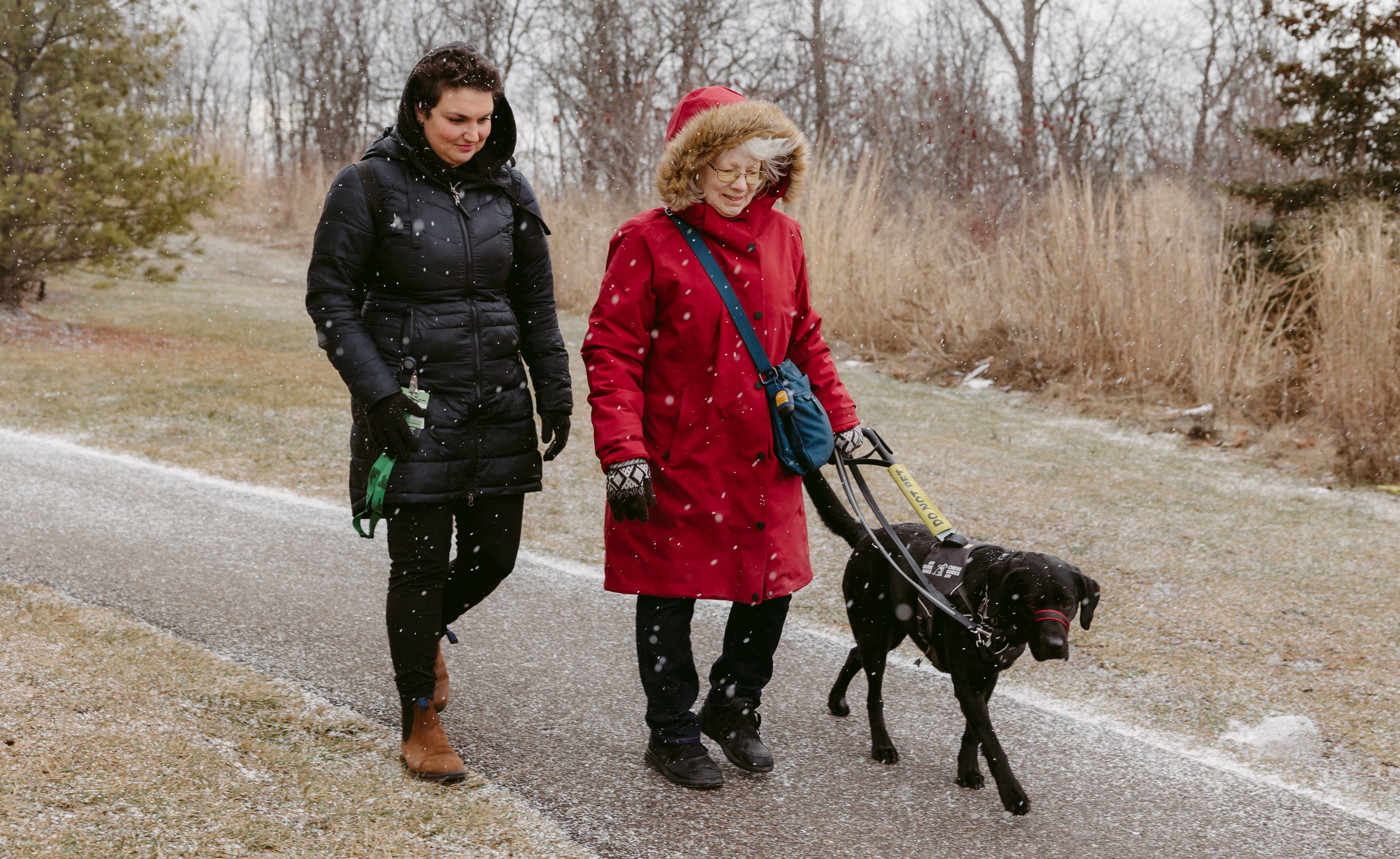 Cheri, who wears a hooded red parka, walks along a paved path with Sassy, her CNIB black Labrador guide dog, and an intervenor from CNIB Deafblind Community Services.