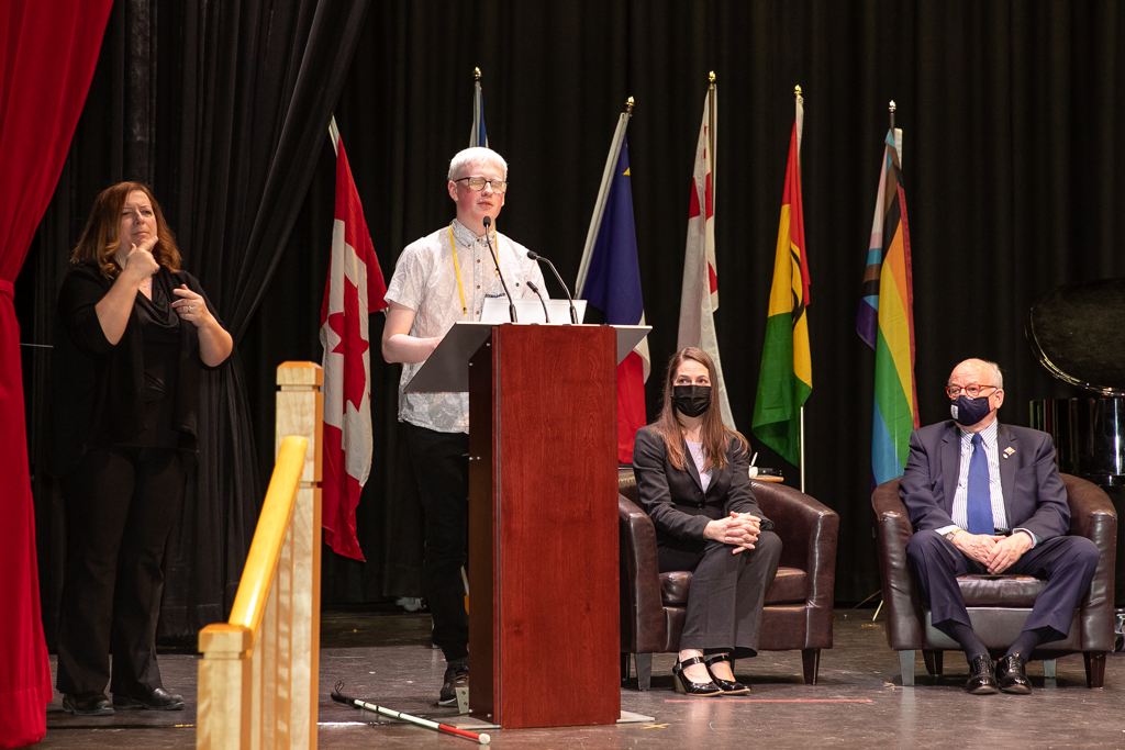Caelin stands at a podium and speaks at the Nova Scotia Education Week Awards ceremony. In the background sits Hon. Arthur J. Leblanc and Hon. Minister Becky Druhan.