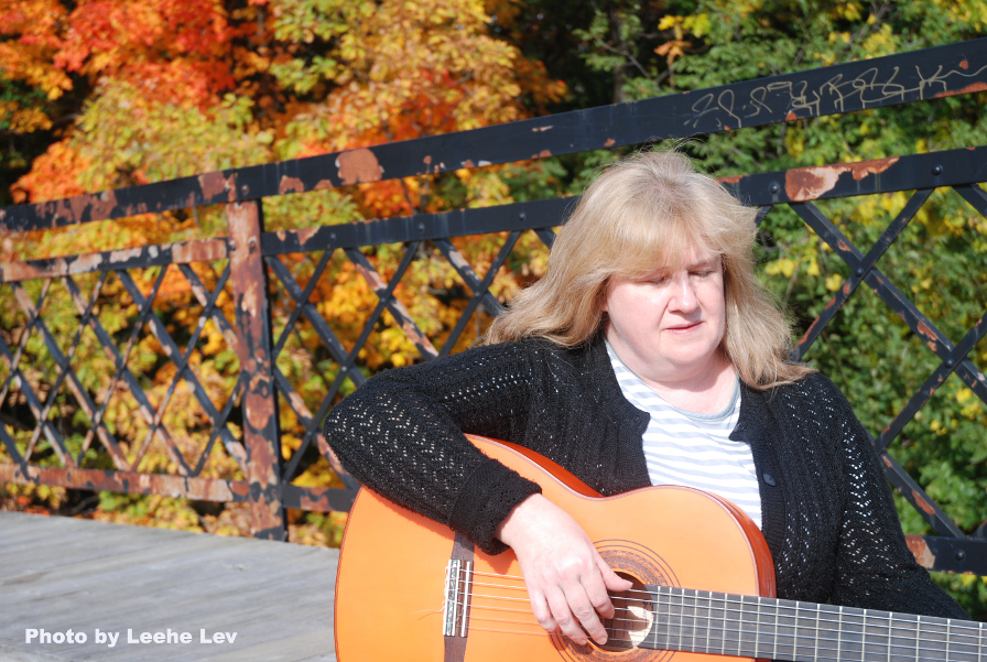 Photo de Christine en train de jouant la guitare sur un pont. Il y a des arbres derrière elle et le soleil brille.