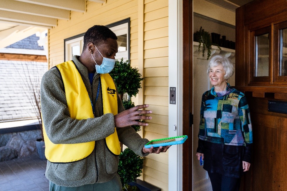 Un collecteur de fonds d'INCA portant un masque et un gilet jaune, tenant une tablette, discutant avec une donatrice sur le pas de sa porte.