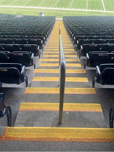 A flight of concrete stadium stairs from the top looking downwards towards a football field, with rows of stadium seats on either side. The stairs are divided by a railing in the middle, and each stair is painted a bright golden yellow along the length of the front edges.