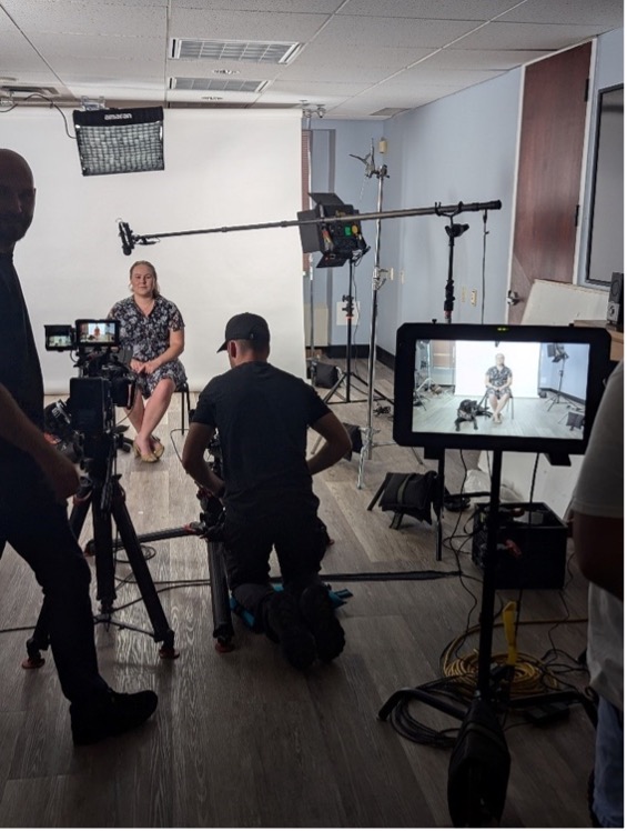 Taylor Bauer and her guide dog, Wallace, are interviewed for a public education video. Taylor sits with a white screen behind her while several production staff operate a camera and recording equipment in front of her.