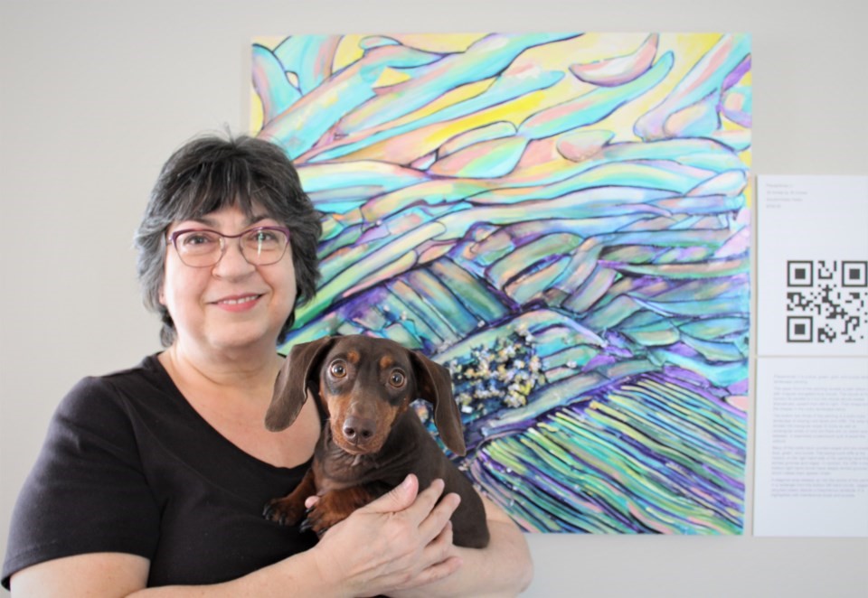 Robyn Rennie pose pour une photo devant son œuvre d'art colorée à côté d'un code QR imprimé numériquement. Elle tient dans ses bras un petit chien brun chocolat. 