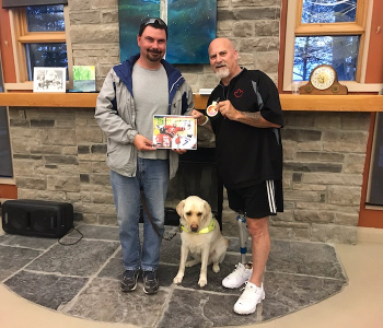 Guy Carrière et son chien-guide (à gauche) et Paul Rosen (à droite) posent ensemble pour une photo. Guy tient une photo autographiée de Paul et Paul montre une médaille dans la main gauche. 