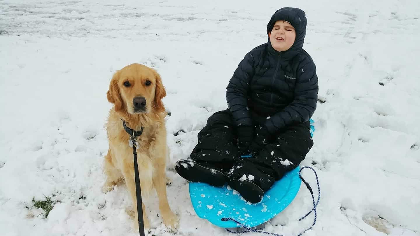 Landon et Ruggles, un Golden Retriever, assis l'un à côté de l'autre dans la neige, tout sourire pour l'appareil photo; Landon en habit de neige est assis sur un traîneau