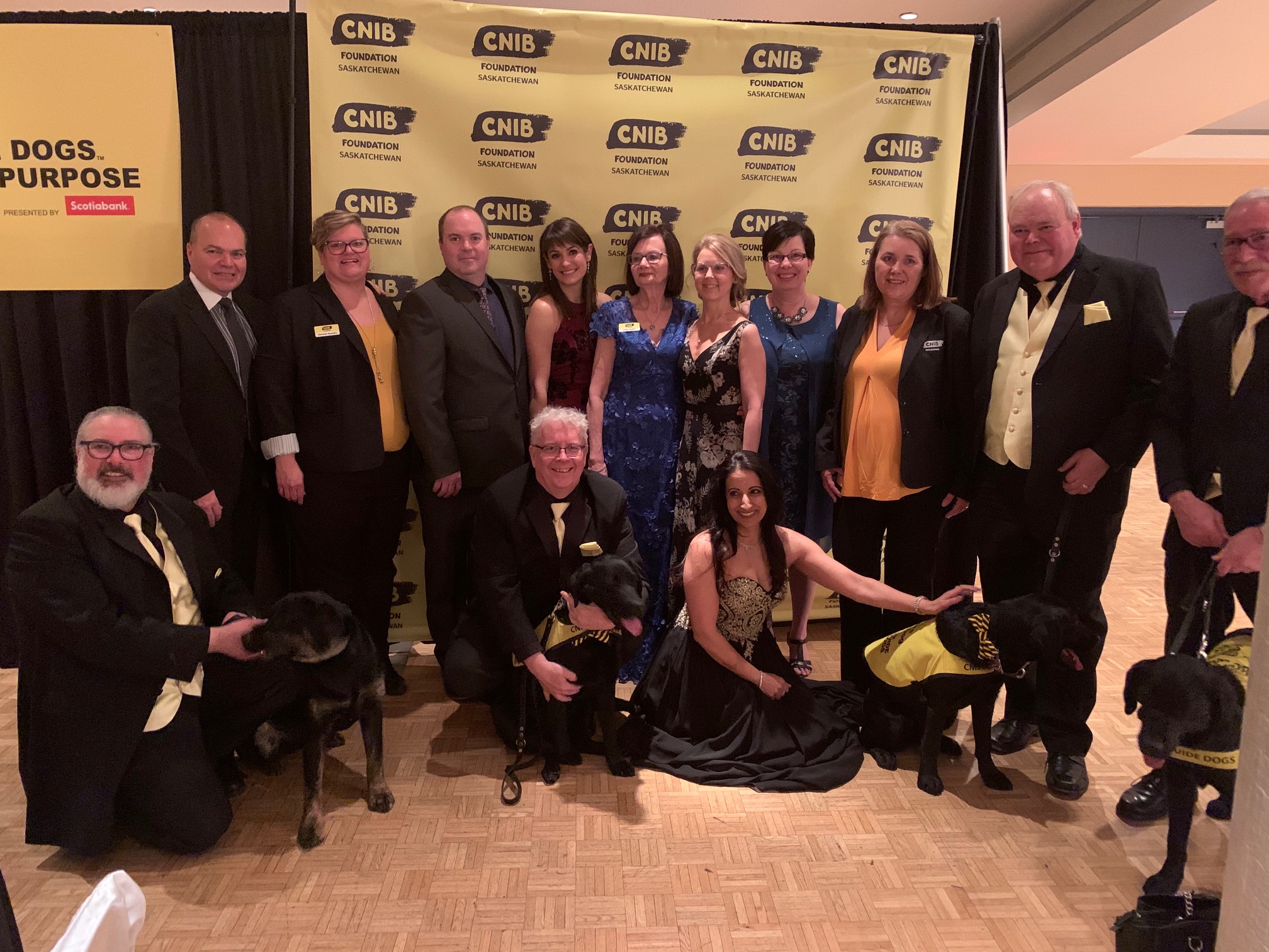 A large group of CNIB Staff and Volunteers pose in front of a branded CNIB wallpaper at the Guide Dogs with Purpose Gala.