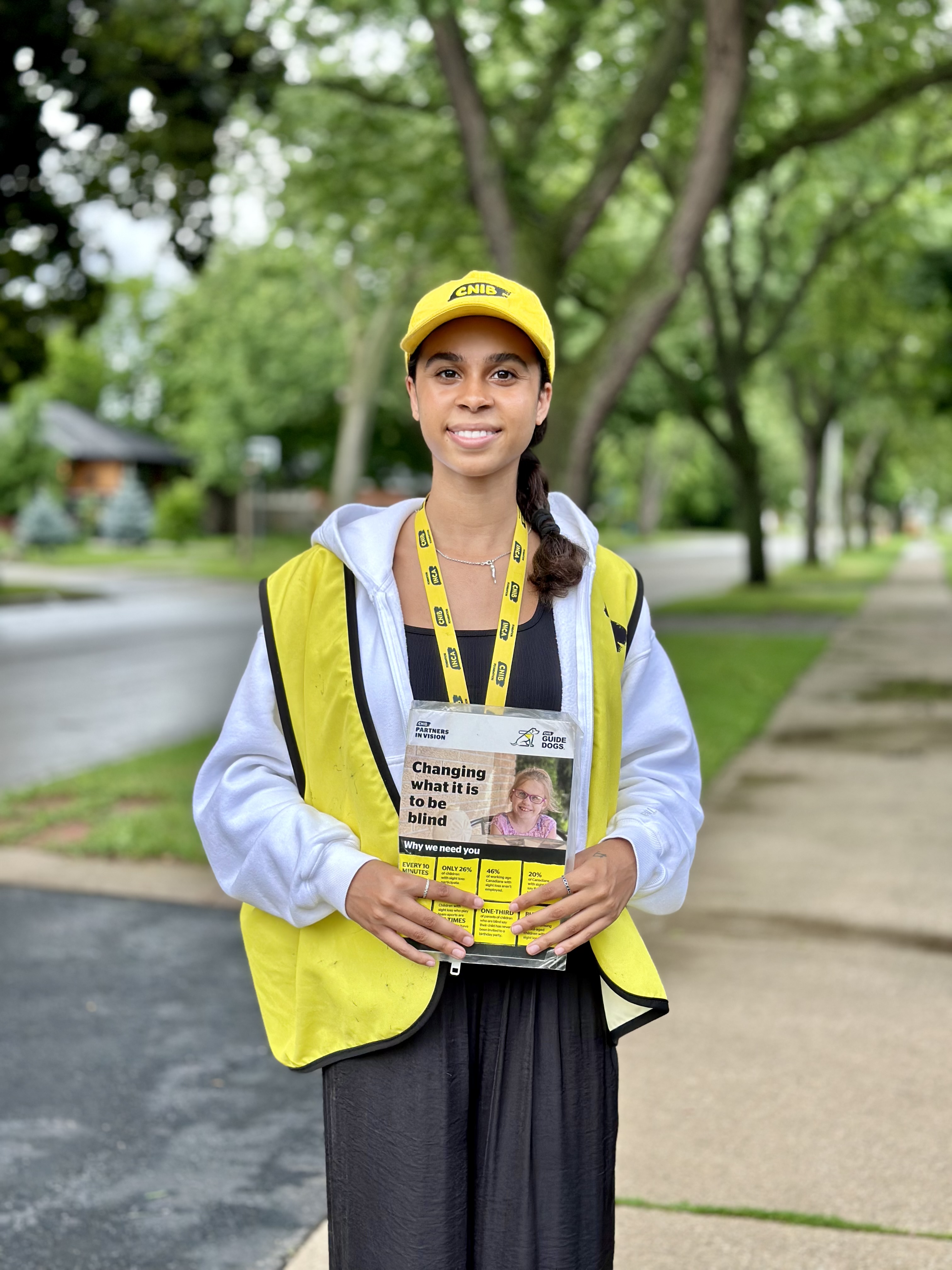 A canvasser working on behalf of CNIB smiles for the camera while standing on a sidewalk underneath some lush trees. The canvasser wears a yellow CNIB hat, lanyard with photo ID, and vest, and holds a large card that features facts about blindness.