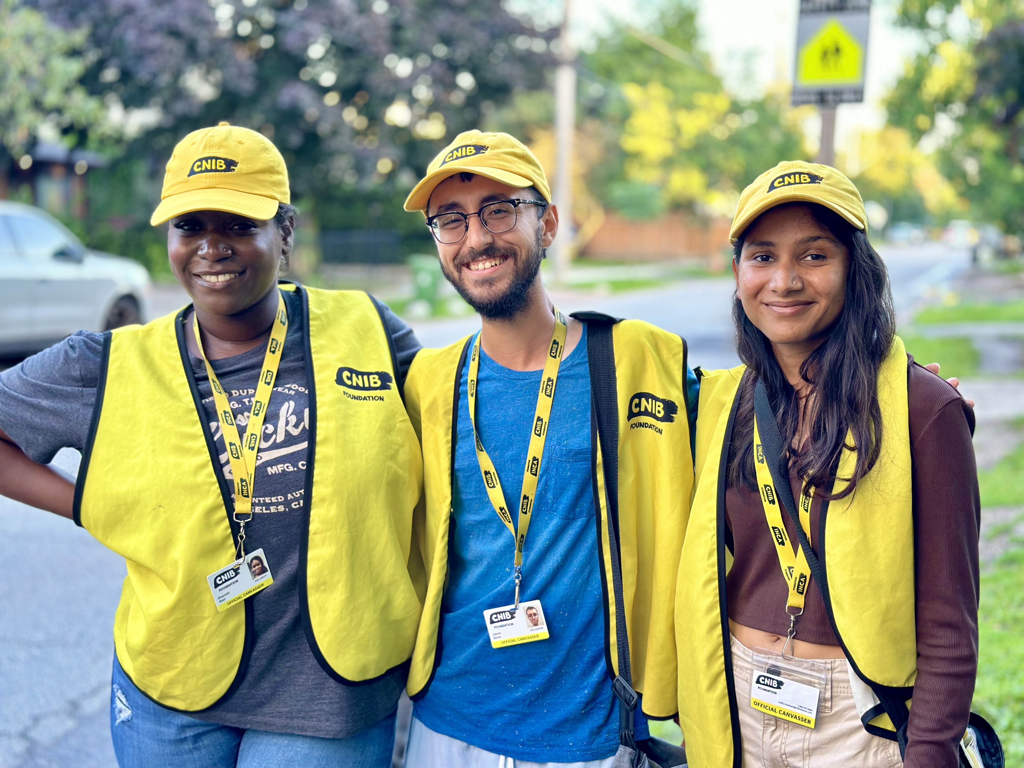 Three canvassers working on behalf of CNIB smile for the camera while standing on a sidewalk in a residential neighbourhood. Each canvasser is wearing a yellow CNIB baseball cap, vest, and lanyard with photo identification.