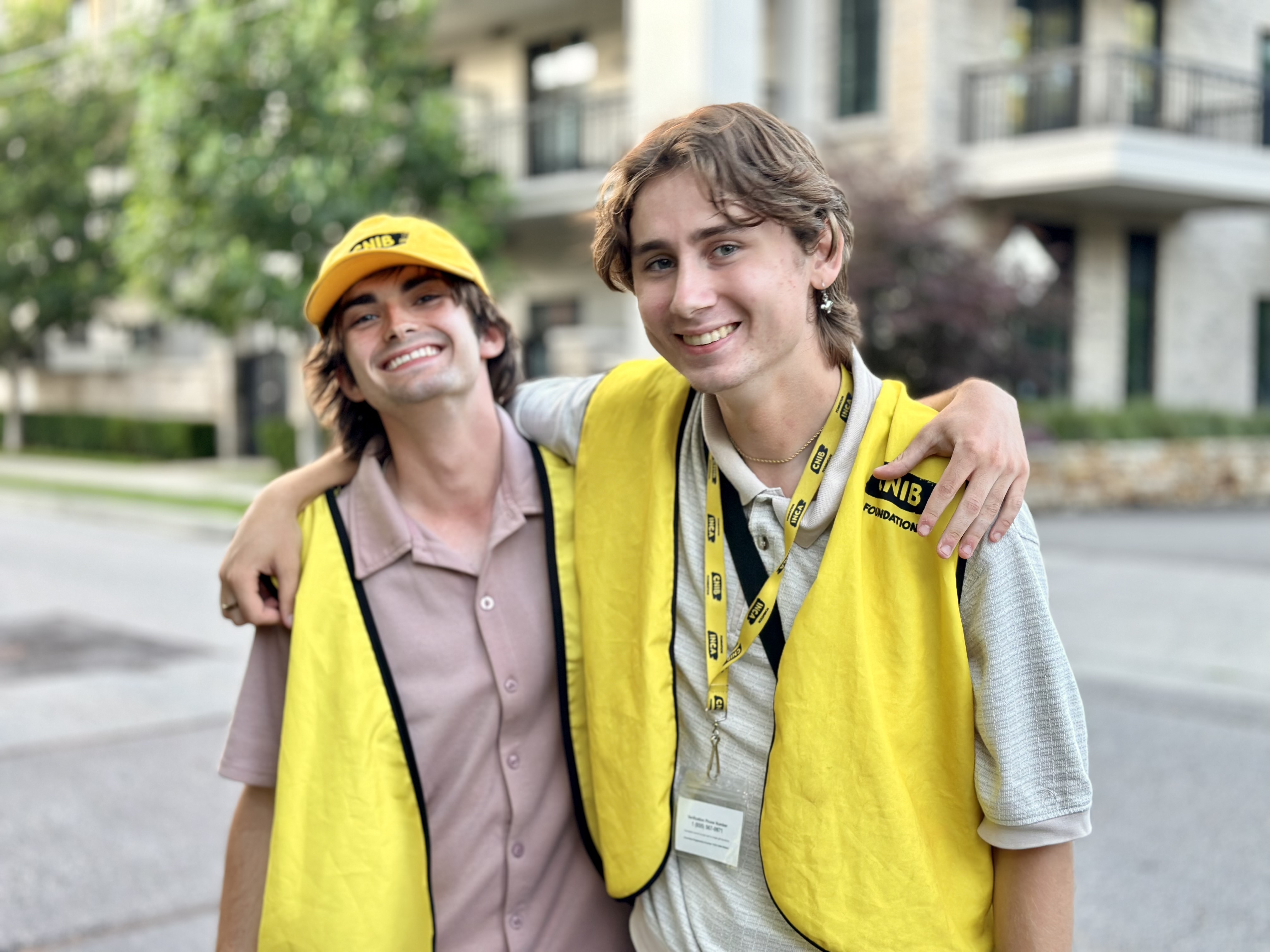 Two canvassers working on behalf of CNIB smile with their arms around each other while standing in front of an apartment building. Both canvassers are wearing a yellow CNIB vest. The canvasser on the left is also wearing a yellow CNIB baseball cap, while the one on the right also wears a yellow lanyard.