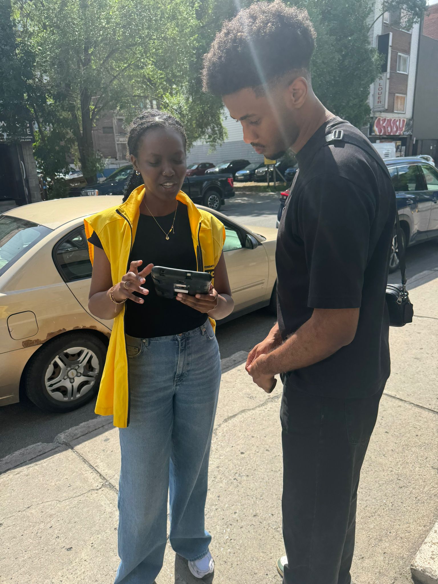 A canvasser working on behalf of CNIB speaks to a person on the sidewalk while both review the screen of her secure tablet device. The canvasser wears a yellow CNIB vest.