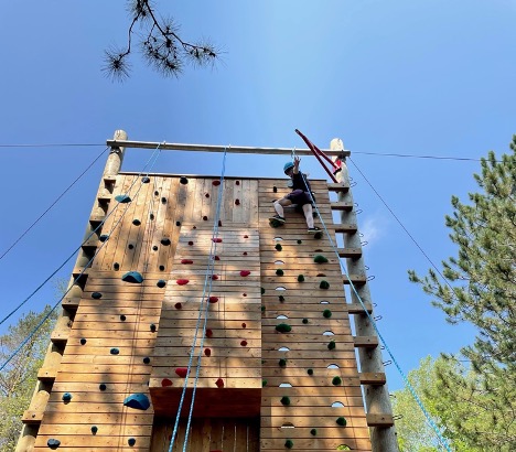 Julia scales the climbing tower at CNIB Lake Joe. She is wearing a harness and helmet. 