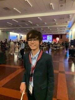 Keaton at the Braille Challenge finalist’s party. He is indoors at a banquet hall and wearing a suit while carrying a white cane in his right hand.