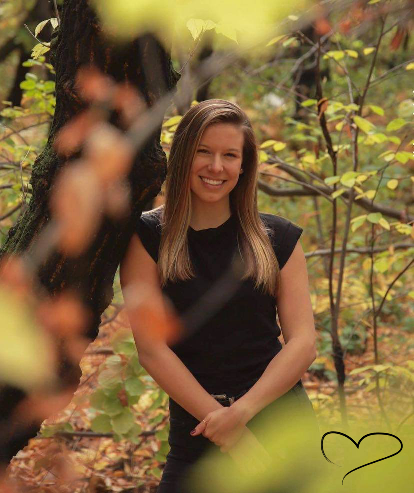 Kimberly Blain, leaning against a tree in a forest during the Autumn season. A heart graphic can be seen in the leaves in the bottom right corner of the picture