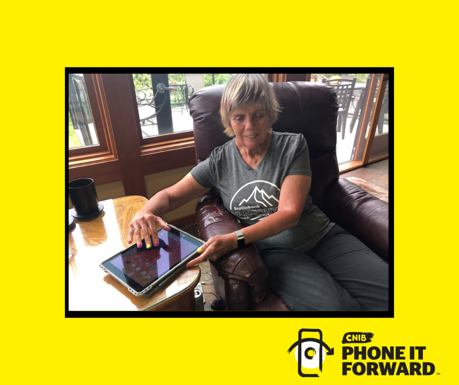 Marilyn sits on a chair and types on an iPad. The iPad is sitting on top of a coffee table to the right of the chair. The photograph appears on a yellow background with a picture frame outline. Phone it Forward logo bottom-right corner.