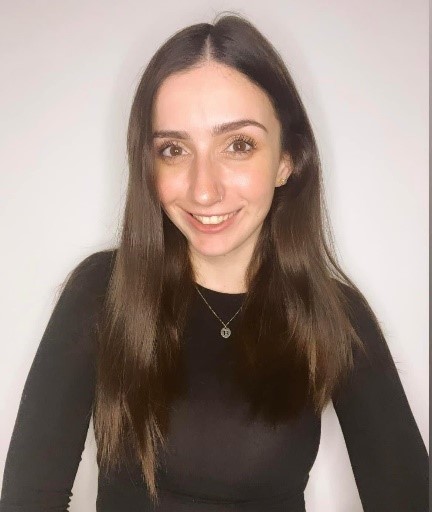 A young woman with long brunette hair is smiling at the camera. She is wearing a long-sleeved black shirt and a necklace.