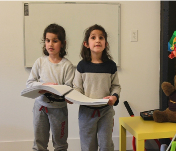 Deux jeunes enfants tiennent un même livre en braille et se promènent dans une salle de classe.]